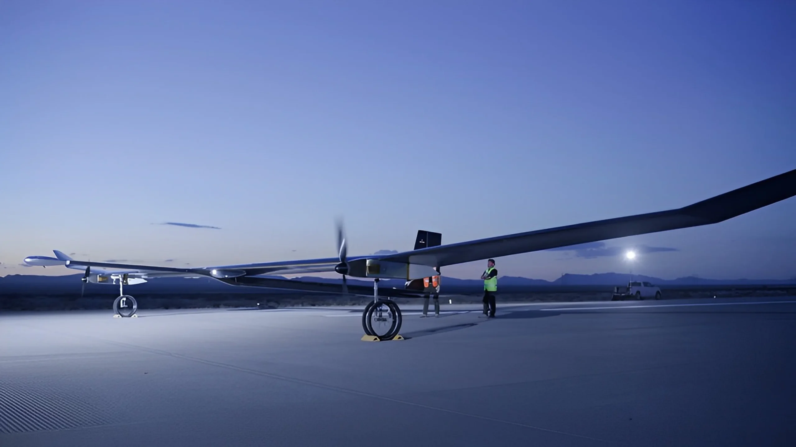 Dawn scene on an airstrip with a long-winged drone preparing for flight, assisted by ground crew under a rising moon.