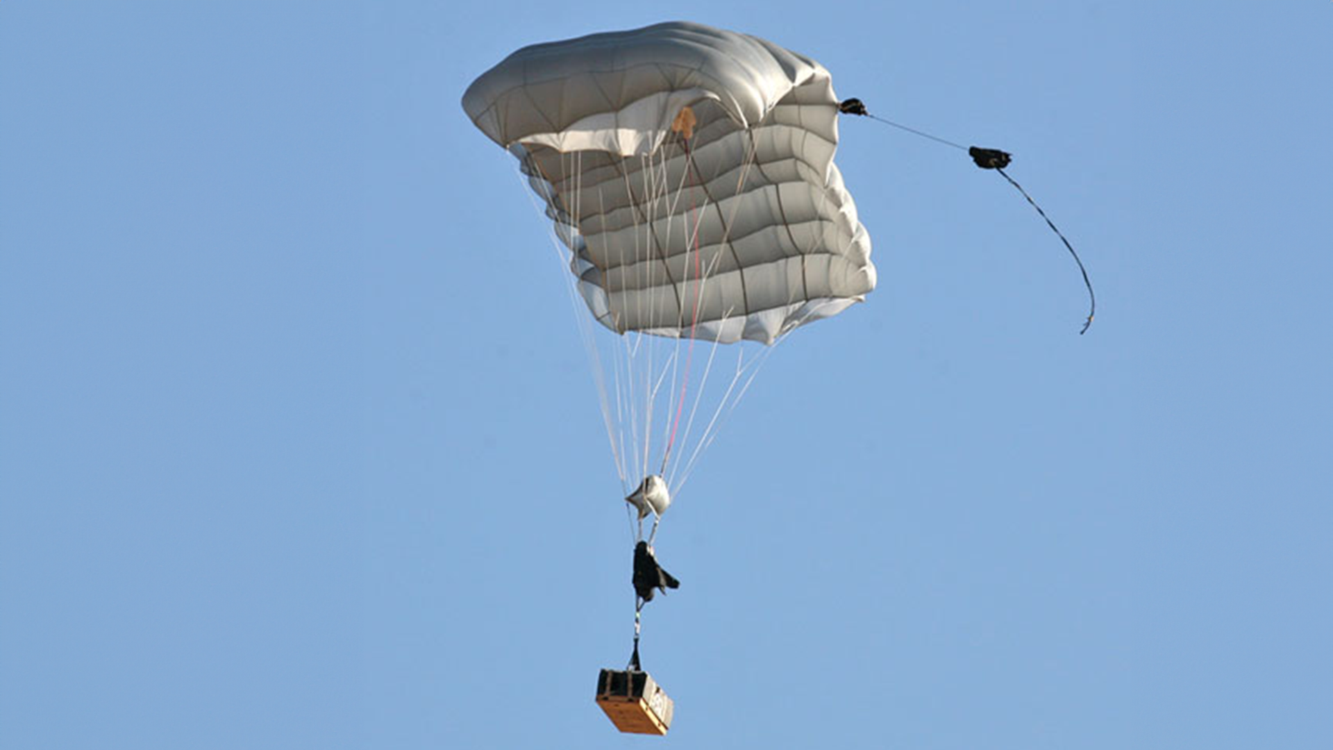 Autonomous parachute system in flight, carrying a cargo box below it, demonstrating the capability for precise delivery in a clear blue sky.