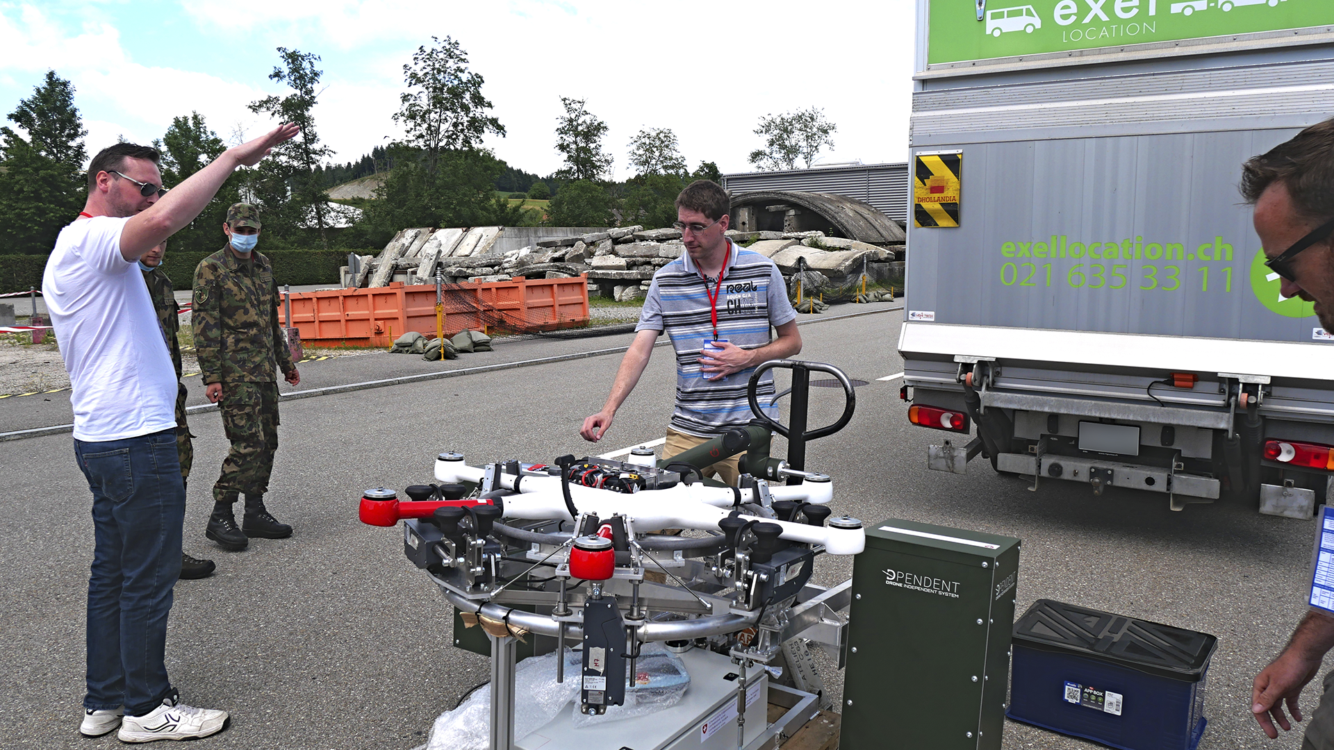 A group of individuals, including a soldier and two men, interact around a drone ground handling station outdoors, while a delivery truck is parked in the background.