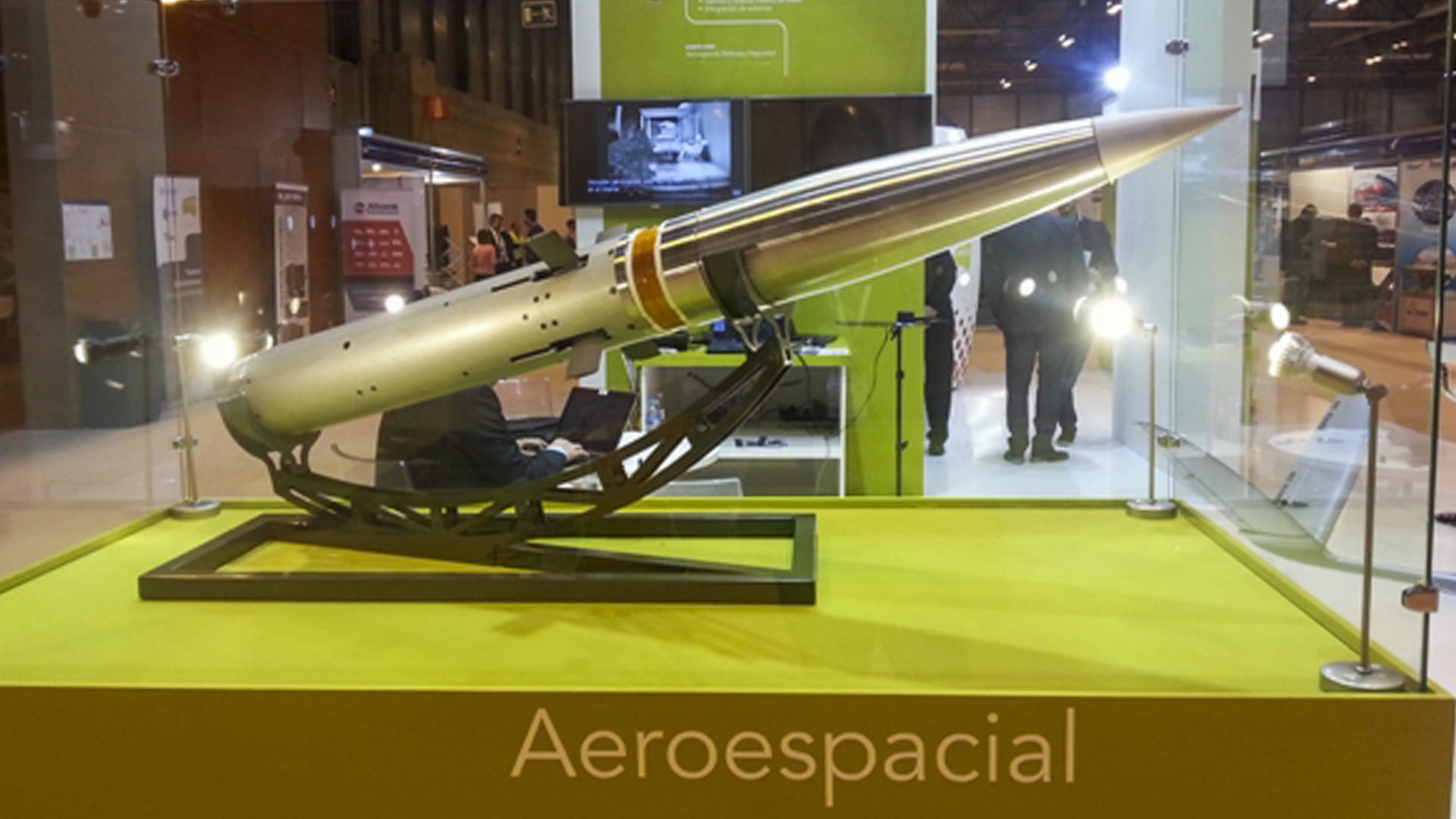 Precision-guided missile on display in a glass case at an exhibition, featuring its sleek metallic design and mounted on a stand, with background activity and signage visible.