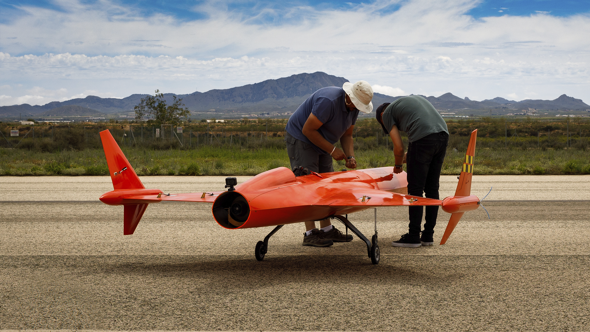 Two technicians inspecting and preparing a bright red unmanned aerial vehicle (UAV) on a runway with mountains in the background.