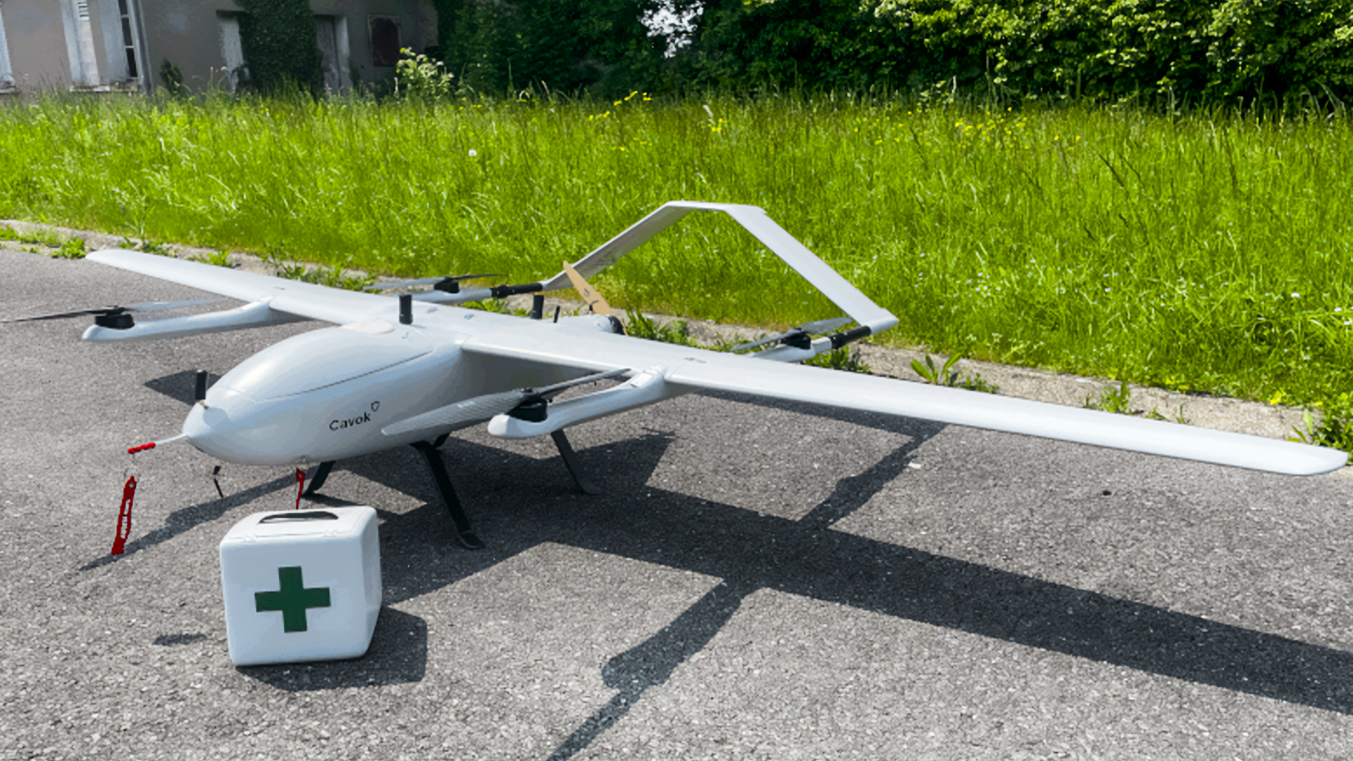 Fixed-wing drone equipped for medical supply delivery, positioned next to a first aid kit on a paved surface, with green grass in the background.