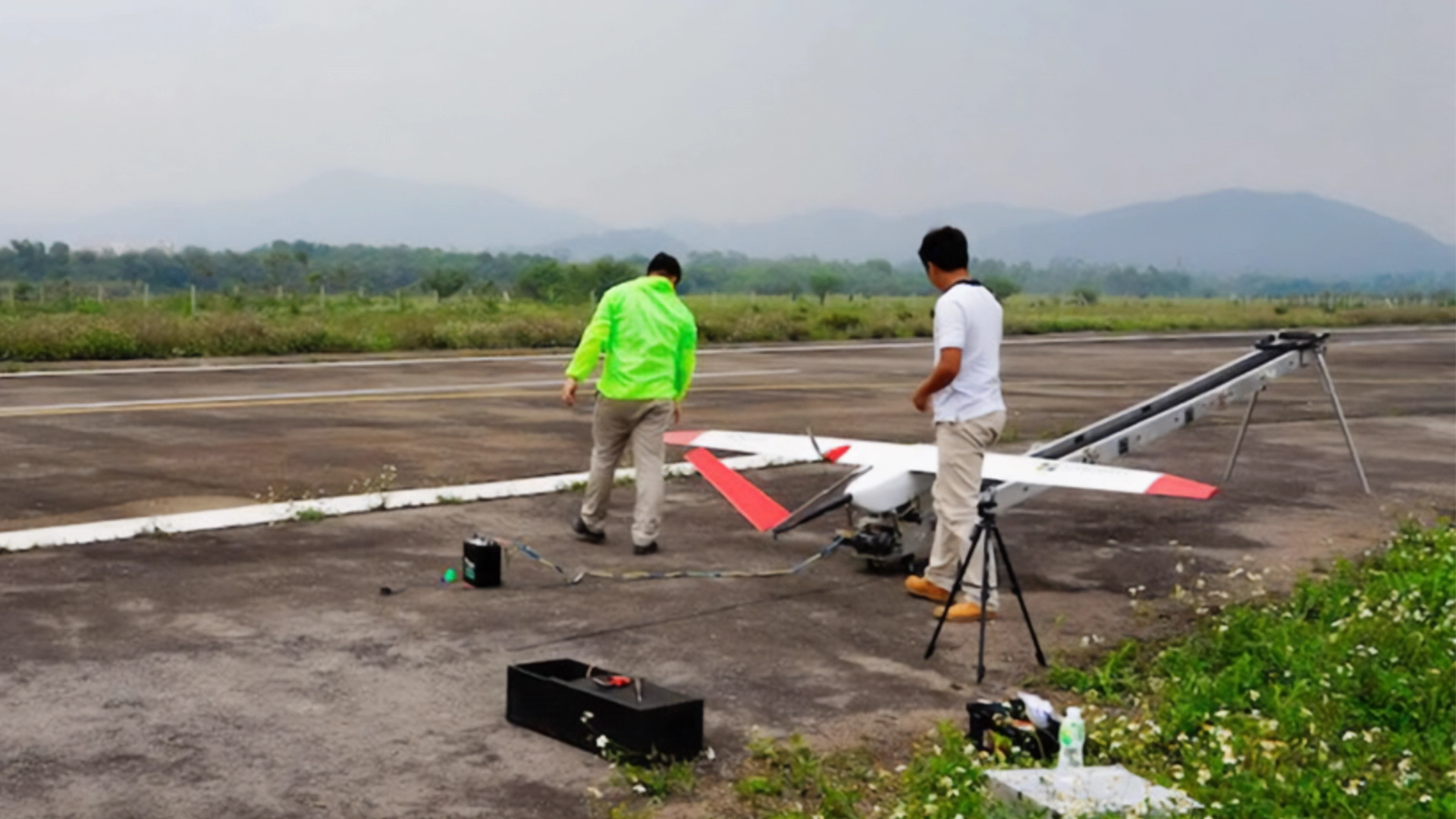 Two technicians preparing the AS-III UAS for launch using a catapult on a runway, with a green landscape in the background.