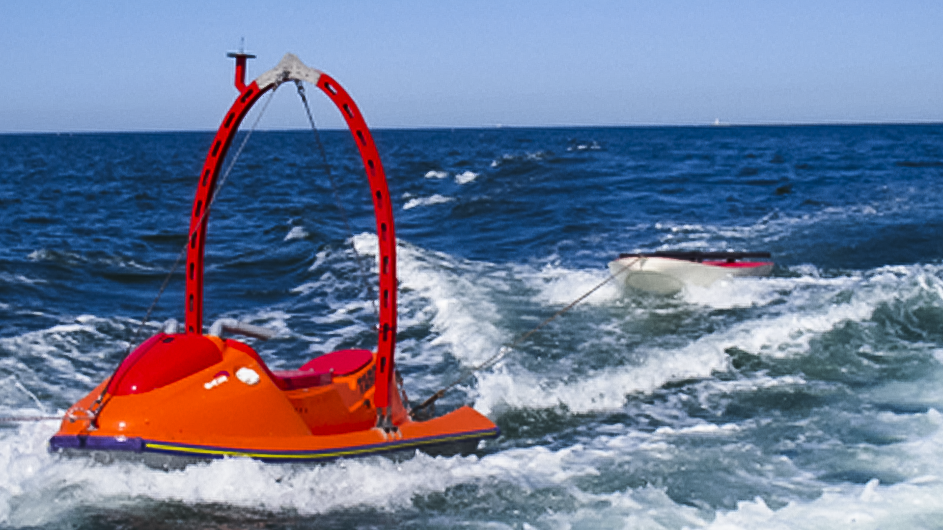 Orange unmanned surface vehicle (USV) navigating through waves at sea, equipped with a red arched structure, towing a target in the background.